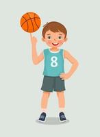 lindo niño pequeño con ropa deportiva girando baloncesto en su dedo posando con la mano en la cintura vector