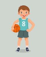 lindo niño pequeño con ropa deportiva sosteniendo baloncesto de pie posando con la mano en la cintura vector