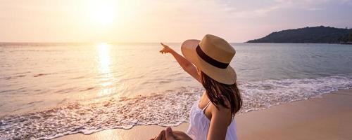Young woman traveler holding man's hand and looking beautiful sunset on the beach, Couple on vacation in summer concept photo