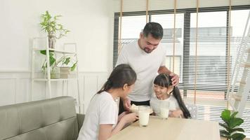 uma família tailandesa asiática saudável, filha e jovem mãe bebem leite branco fresco em vidro e pão do pai, alegria juntos em uma mesa de jantar de manhã, bem-estar nutrição em casa café da manhã refeição estilo de vida. video