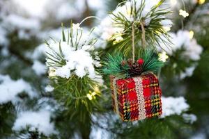 Christmas gift box on a branch of a natural spruce covered with snow. New year outdoor. Snowfall, garland lights, festive mood of fairy tales and magic, street decoration. Copy space photo