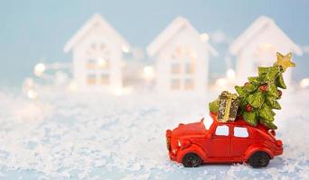 Christmas decor - red retro car on snow carries past white houses with lights garlands in bokeh Christmas tree with gift boxes on roof. Toy on blue background, space for text. New Year in town photo