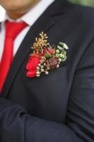 The groom's wedding boutonniere of succulents and red flowers in a black jacket with a red tie. Festive decor, flowers, attire for the marriage registration. Close-up, space for text photo