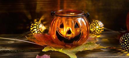Lamp pumpkin with eyes and mouth made of glass and natural orange pumpkin on a wooden table with yellow and red maple leaves. Halloween, warm autumn atmosphere. Round garland, candle in a candlestick. photo