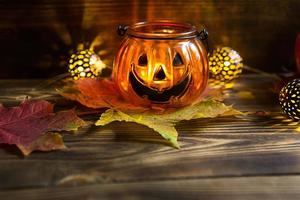 Lamp pumpkin with eyes and mouth made of glass and natural orange pumpkin on a wooden table with yellow and red maple leaves. Halloween, warm autumn atmosphere. Round garland, candle in a candlestick. photo