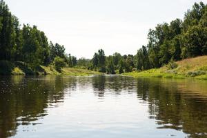 río en verano en un día soleado, hierba verde y bosque. reflejo en el agua. turismo interno, ecoturismo, unidad con la naturaleza. medio ambiente limpio, protección. caminatas acuáticas y campamentos. Hora de verano. foto