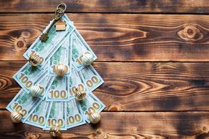 Christmas tree made of 100 dollar bills on wooden background with copyspace and House key. Christmas decor of finance, savings, wealth, expenses in new year. Real estate, housing, mortgage, relocation photo