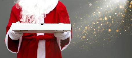 White pizza box in hands of Santa Claus in mittens, with a beard, in a red coat. Christmas fast food delivery. New year's eve promotion. Work on public holidays catering. Copy space, mock up. Banner photo