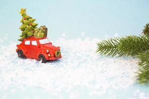 Christmas decor - a red retro car on artificial snow carries a Christmas tree with gift boxes on the roof. Toy with sequins on a blue background, space for text. New Year photo