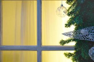 Christmas decorations made of spruce branches and white transparent balls and ribbons frame the window with wooden frame and yellow light. New year festive atmosphere, comfort of home. Space for text photo