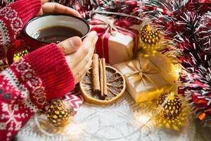 la mano de una mujer en un suéter cálido sostiene una taza roja con una bebida caliente en una mesa con adornos navideños. ambiente de año nuevo, palitos de canela y una rodaja de naranja seca, regalos, guirnaldas y oropel foto