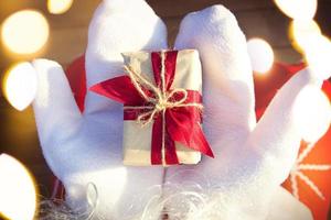 A box with a Christmas gift in the hands of Santa Claus in white mittens. Red suit, beard, garland lights in a blur. New year, preparation, expectation of a miracle, a dream come true. Close up photo