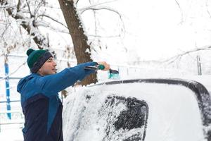 A man brushes snow from a car after a snowfall. A hand in a blue jacket with a car broom on the white body. Winter weather conditions photo