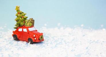 Christmas decor - a red retro car on artificial snow carries a Christmas tree with gift boxes on the roof. Toy with sequins on a blue background, space for text. New Year photo