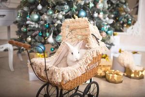 un conejo blanco se sienta dentro de un cochecito de bebé retro para muñecas. decoración navideña, árbol de navidad con guirnaldas de luces. Año Nuevo. mascotas en casa foto