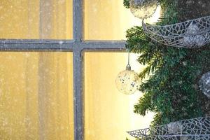 Christmas decorations made of spruce branches and white transparent balls and ribbons frame the window with wooden frame and yellow light. New year festive atmosphere, comfort of home. Space for text photo