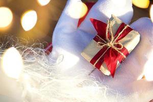 una caja con un regalo de navidad en manos de santa claus en mitones blancos. traje rojo, barba, guirnaldas de luces borrosas. año nuevo, preparación, expectativa de un milagro, un sueño hecho realidad. de cerca foto