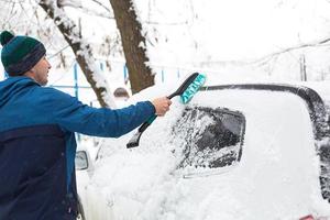 A man brushes snow from a car after a snowfall. A hand in a blue jacket with a car broom on the white body. Winter weather conditions photo