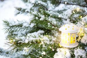 Christmas lantern with windows on branch of natural spruce in snow. New year outdoor. Snowfall, garland lights, festive mood of fairy tales and magic, street decoration. Copy space photo