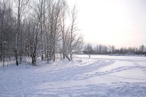Winter landscape - white snow with traces of shoes and skis on the field. The ski track and road skirting the forest with bare trees, soft sunlight. photo