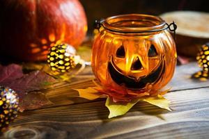 Lamp pumpkin with eyes and mouth made of glass and natural orange pumpkin on a wooden table with yellow and red maple leaves. Halloween, warm autumn atmosphere. Round garland, candle in a candlestick. photo