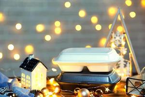 Christmas decor of food delivery service containers. New year's eve promotion. Ready-made hot order, disposable plastic box in fairy light. Work on public holidays catering. Copy space, mock up photo