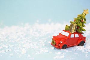 Christmas decor - a red retro car on artificial snow carries a Christmas tree with gift boxes on the roof. Toy with sequins on a blue background, space for text. New Year photo