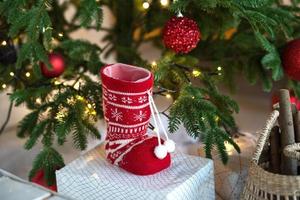 Red knitted sock with ornaments - rustic Christmas decor under the Christmas tree with lights of garlands. New year, home decor elements, comfort, warmth. photo