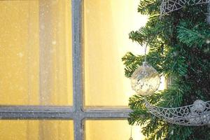 Christmas decorations made of spruce branches and white transparent balls and ribbons frame the window with wooden frame and yellow light. New year festive atmosphere, comfort of home. Space for text photo