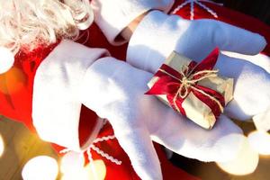 A box with a Christmas gift in the hands of Santa Claus in white mittens. Red suit, beard, garland lights in a blur. New year, preparation, expectation of a miracle, a dream come true. Close up photo