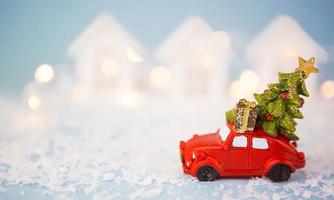 Christmas decor - red retro car on snow carries past white houses with lights garlands in bokeh Christmas tree with gift boxes on roof. Toy on blue background, space for text. New Year in town photo