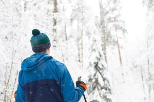 Skier in windbreaker and hat with pompom with ski poles in his hands with his back against the background of a snowy forest. Cross-country skiing in winter forest, outdoor sports, healthy lifestyle. photo