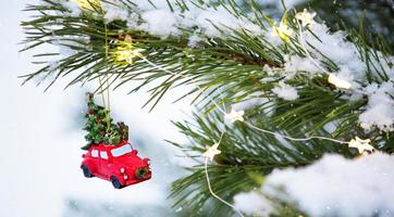 Red Christmas toy retro car with a Christmas tree on branch of natural spruce in snow. New year outdoor. Snowfall, garland lights, festive mood of fairy tales and magic, street decoration. Copy space photo