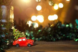 Christmas decor - red retro car carries Christmas tree with gift boxes on roof. space for text. New Year. Toy on branch of fir tree with Golden lights of garlands in defocus, bokeh, holiday background photo