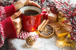 la mano de una mujer en un suéter cálido sostiene una taza roja con una bebida caliente en una mesa con adornos navideños. ambiente de año nuevo, palitos de canela y una rodaja de naranja seca, regalos, guirnaldas y oropel foto