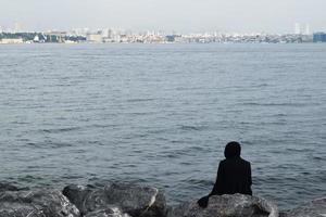 una mujer turca solitaria con un hiyab negro se sienta en la orilla del mar de marmara y mira a estambul foto