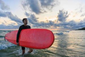 joven surfista asiático sosteniendo una tabla y caminando de regreso a la orilla después de una práctica de deportes acuáticos extremos. foto