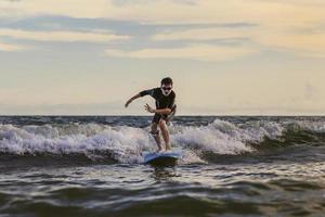 joven niño surfista montando olas con una tabla blanda en la playa de rayong, tailandia. estudiante novato de tablas de surf jugando en el agua con cara emocionada y acción divertida. foto