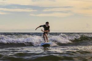 Young boy kid surfer riding waves with a soft board in Rayong beach, Thailand. Rookie surfboard student playing on water in excited face and funny action. photo