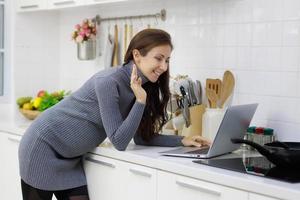 Beautiful, cool, pregnant woman standing in modern kitchen and using laptop computer to chat in videocall with happy and relaxing manner photo