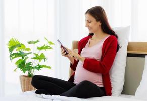 una hermosa mujer caucásica embarazada está felizmente usando su teléfono inteligente en la cama mientras descansa. foto