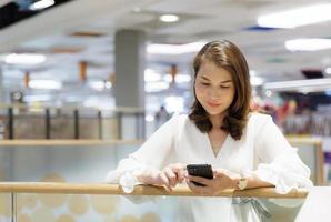 Beautiful Asian mature woman sitting indoors and using smartphone texting and sending while waiting for someone. Attractive female smiles and happy while looking at mobile phone screen photo