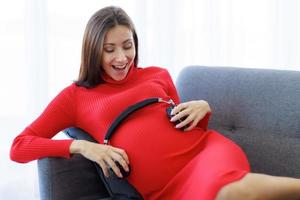 hermosa mujer con una gran mujer embarazada se sienta en el sofá, usa auriculares para sostener su estómago para que el bebé por nacer escuche música.