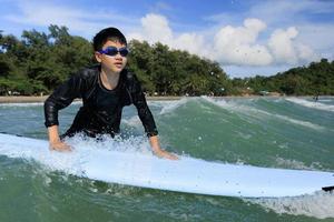 Young boy, student in surfing, is holding onto softboard and trying to bring it back into sea to practice while playing against waves and splashing water. photo