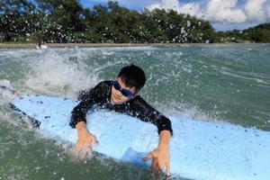 un niño, un estudiante de primer año en el surf, se aferra a la tabla blanda y trata de traerlo de vuelta al mar para practicar mientras juega contra las olas y salpica agua. foto