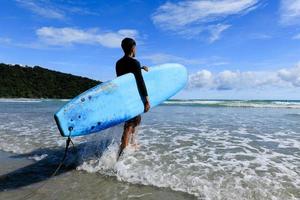 Young fit and firm body teenage guy walking into sea holding soft board ready for extreme sports surfing on beach holidays on clear day, blue skies. photo