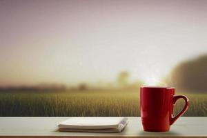 a red cup of coffee and note pad on wooden table and meadows at morning time blurry background photo