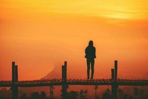 silueta de mujer parada en un puente de madera en la montaña de la colina y el fondo del cielo amarillo al atardecer foto