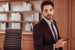 retrato de un joven hombre de negocios inteligente que usa un teléfono inteligente en la sala de trabajo. apuesto hombre de negocios árabe con traje negro sentado y sosteniendo un teléfono inteligente para trabajar. foto