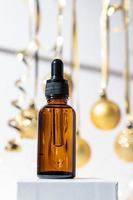 Christmas cosmetics. Pipette dropper bottle made of dark glass with beauty face oil on a sparkling golden baubles background on a white podium. Face and body treatment gift concept photo